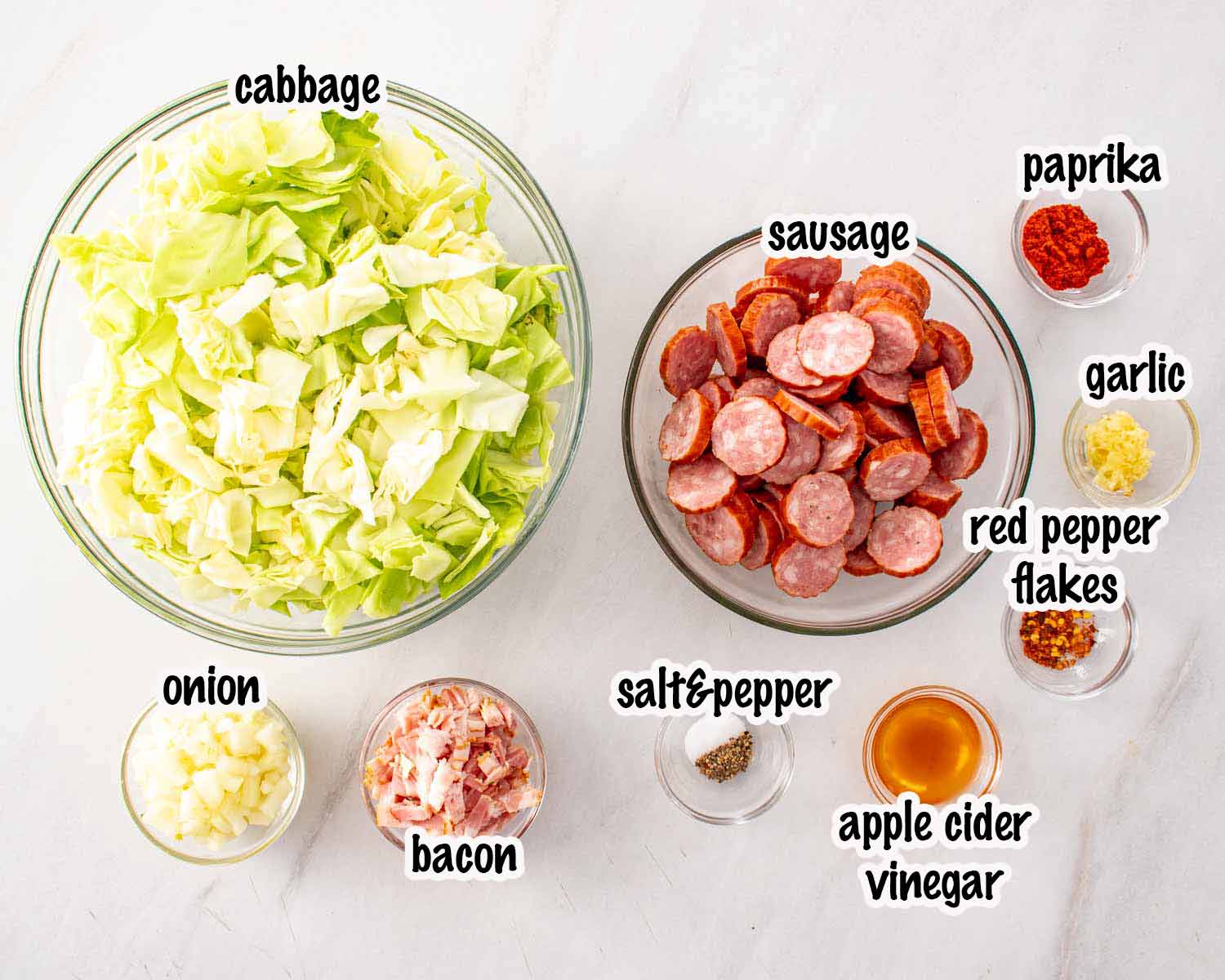 Ingredients for fried cabbage with bacon and sausage arranged in bowls, including cabbage, sausage, bacon, and seasonings.