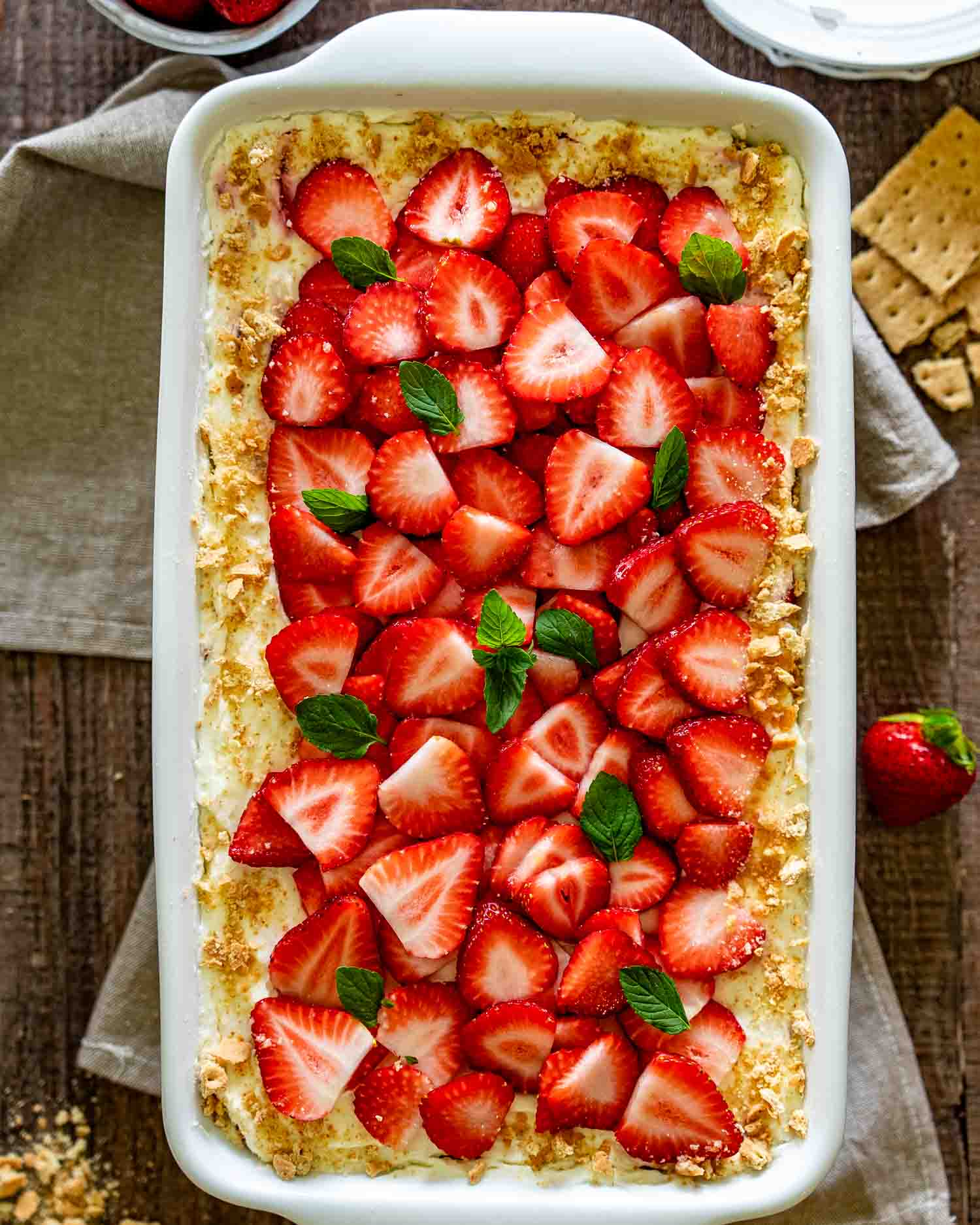 Overhead view of creamy strawberry icebox cake in white baking dish topped with sliced strawberries, mint, and graham cracker crumbs.