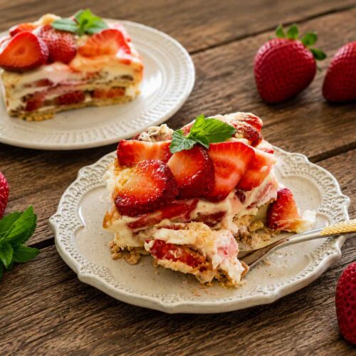 Rustic dessert scene with creamy strawberry icebox cake slices, fresh strawberries, mint leaves, and decorative vintage serving plates.