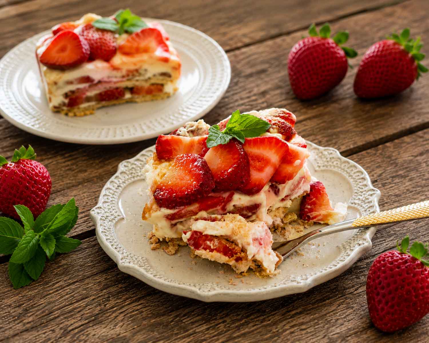 Rustic dessert scene with creamy strawberry icebox cake slices, fresh strawberries, mint leaves, and decorative vintage serving plates.