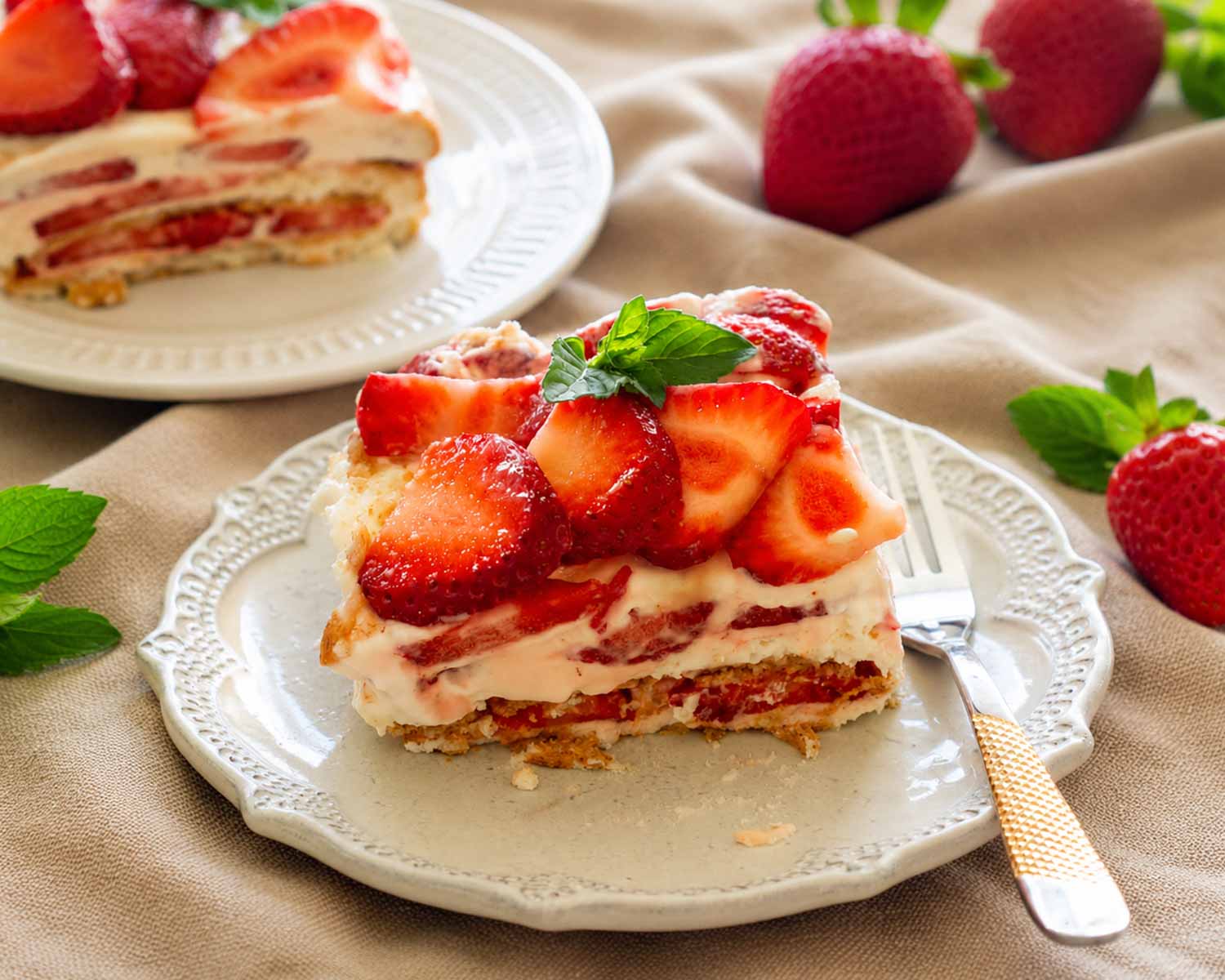 Creamy layered strawberry icebox cake slice topped with fresh strawberries and mint beside gold handled dessert fork on plate.