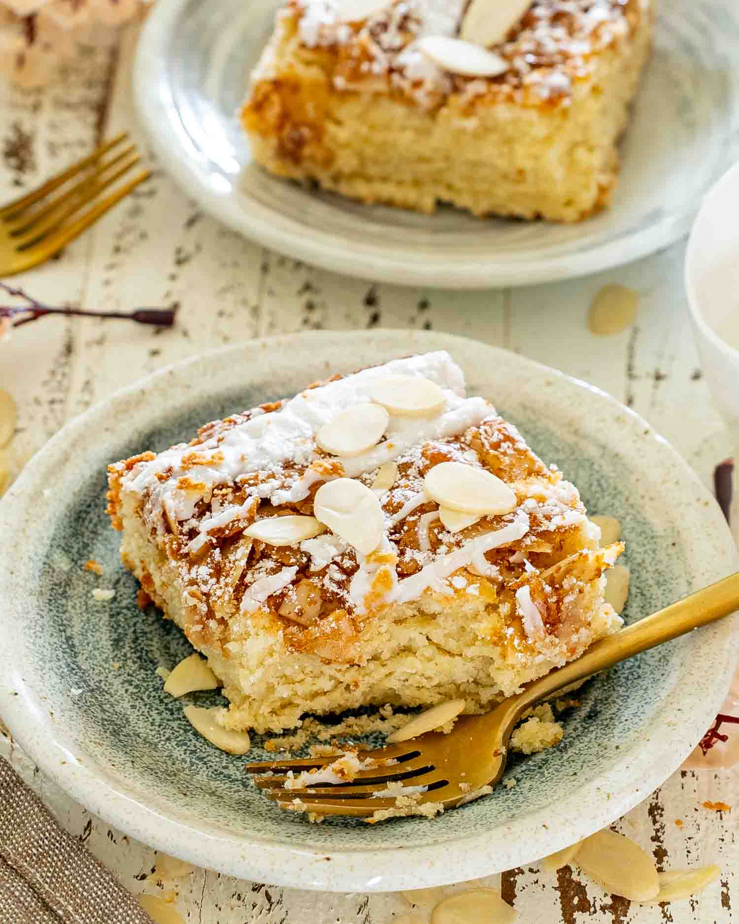 Single almond croissant bar plated with sliced almonds, powdered sugar, icing drizzle, and a warm cup ready for serving.