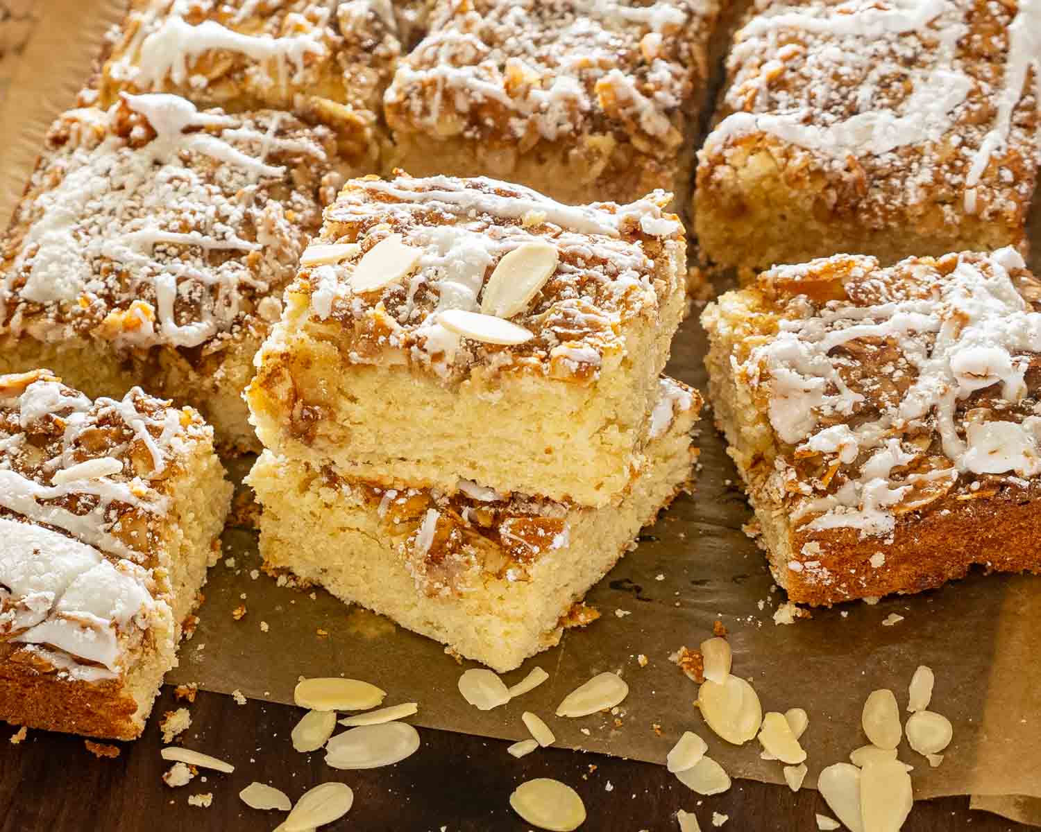 Stacked almond croissant bars showcasing their tender buttery crumb, almond filling, icing drizzle, powdered sugar, and sliced almond topping.