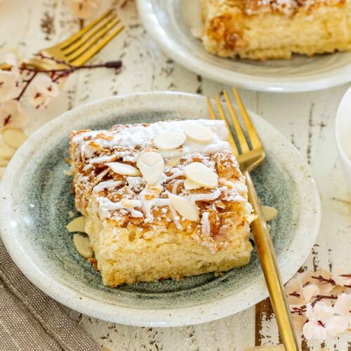 Single almond croissant bar served on a rustic plate with sliced almonds, powdered sugar, icing drizzle, and a gold dessert fork.
