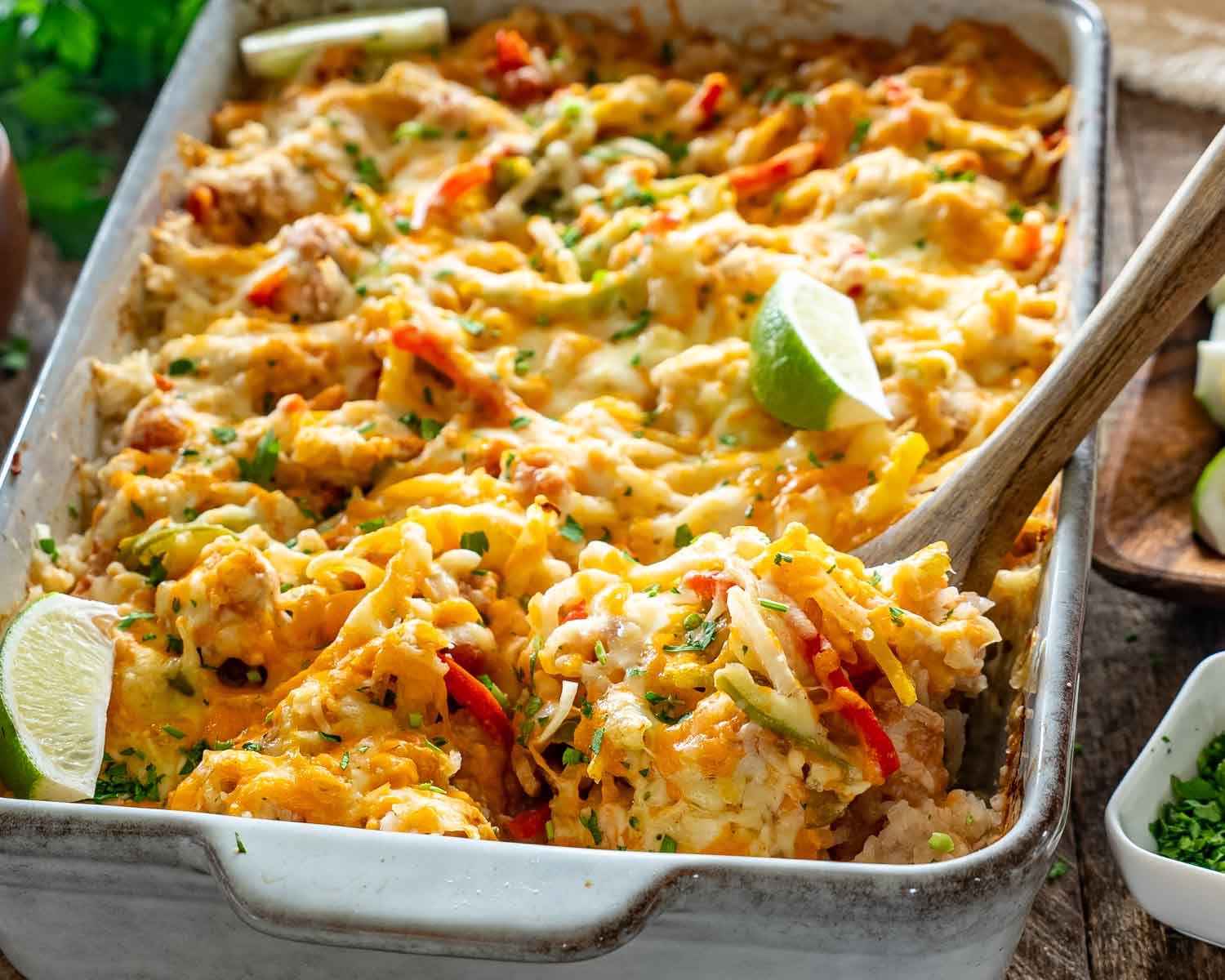 Wooden spoon serving cheesy chicken fajita rice casserole with colorful peppers, melted cheese, cilantro, and fresh lime wedges.