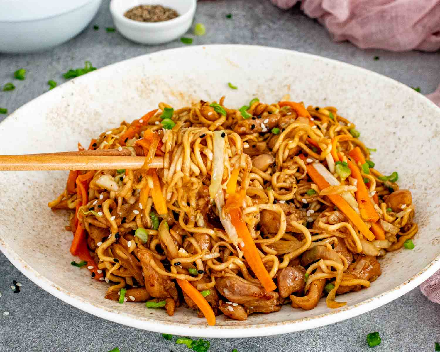 Chopsticks lifting glossy chicken noodle stir fry from a shallow bowl with tender chicken, cabbage, carrots, and sesame seeds.