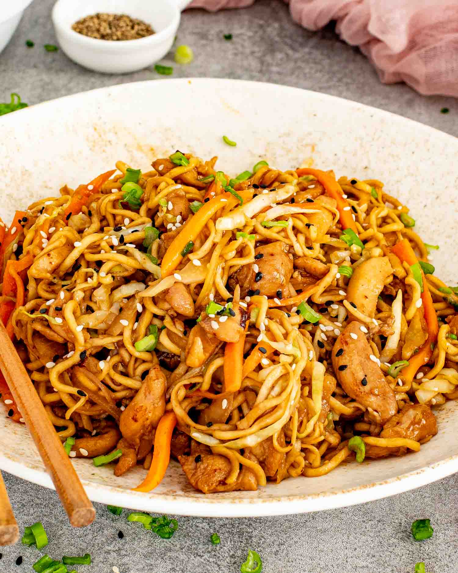 Bowl of homemade chicken noodle stir fry garnished with sesame seeds and green onions, surrounded by garlic, chopsticks, and serving bowls.