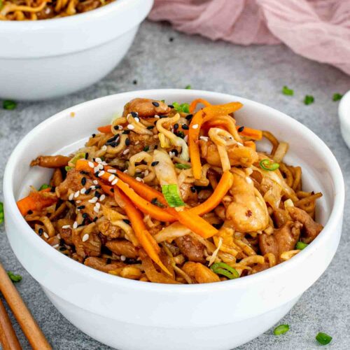 White bowl filled with chicken noodle stir fry garnished with sesame seeds and green onions, with extra bowls blurred behind.