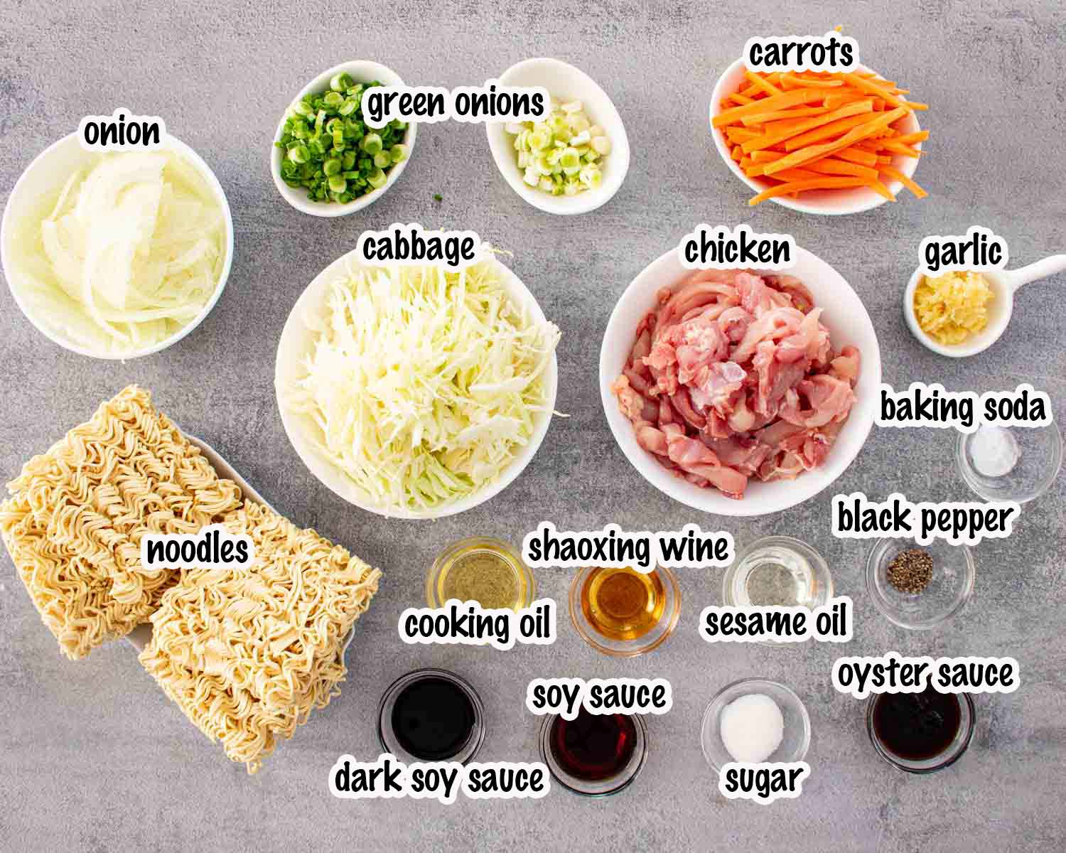 Overhead view of ingredients for chicken noodle stir fry including chicken, ramen noodles, cabbage, carrots, sauces, garlic, and green onions.