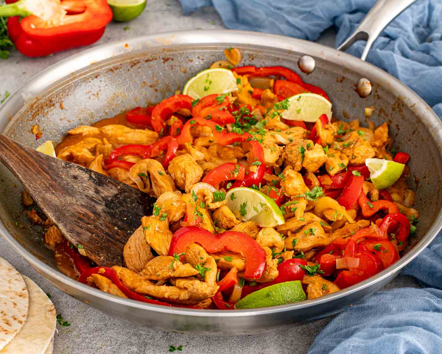 Skillet filled with restaurant style chicken fajitas, red bell peppers, onions, parsley, lime wedges, and a wooden spatula.