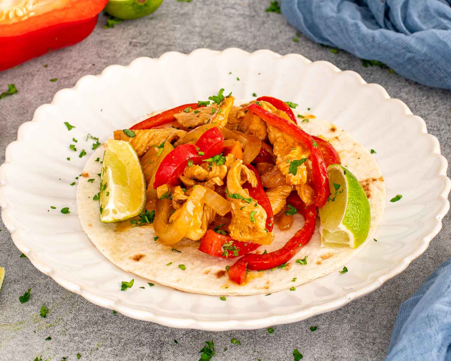 Open flour tortilla topped with seasoned chicken fajitas, sautéed red peppers and onions, parsley, and two fresh lime wedges.