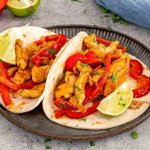 Two soft tortillas piled with saucy chicken fajitas, red bell peppers, onions, parsley, and bright wedges of fresh lime.