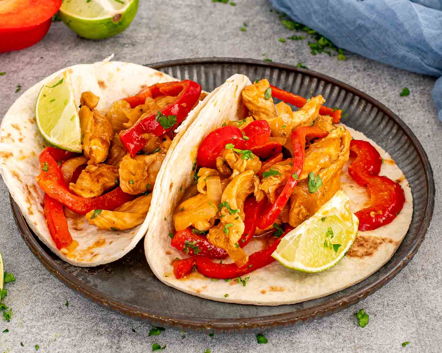 Two soft tortillas piled with saucy chicken fajitas, red bell peppers, onions, parsley, and bright wedges of fresh lime.