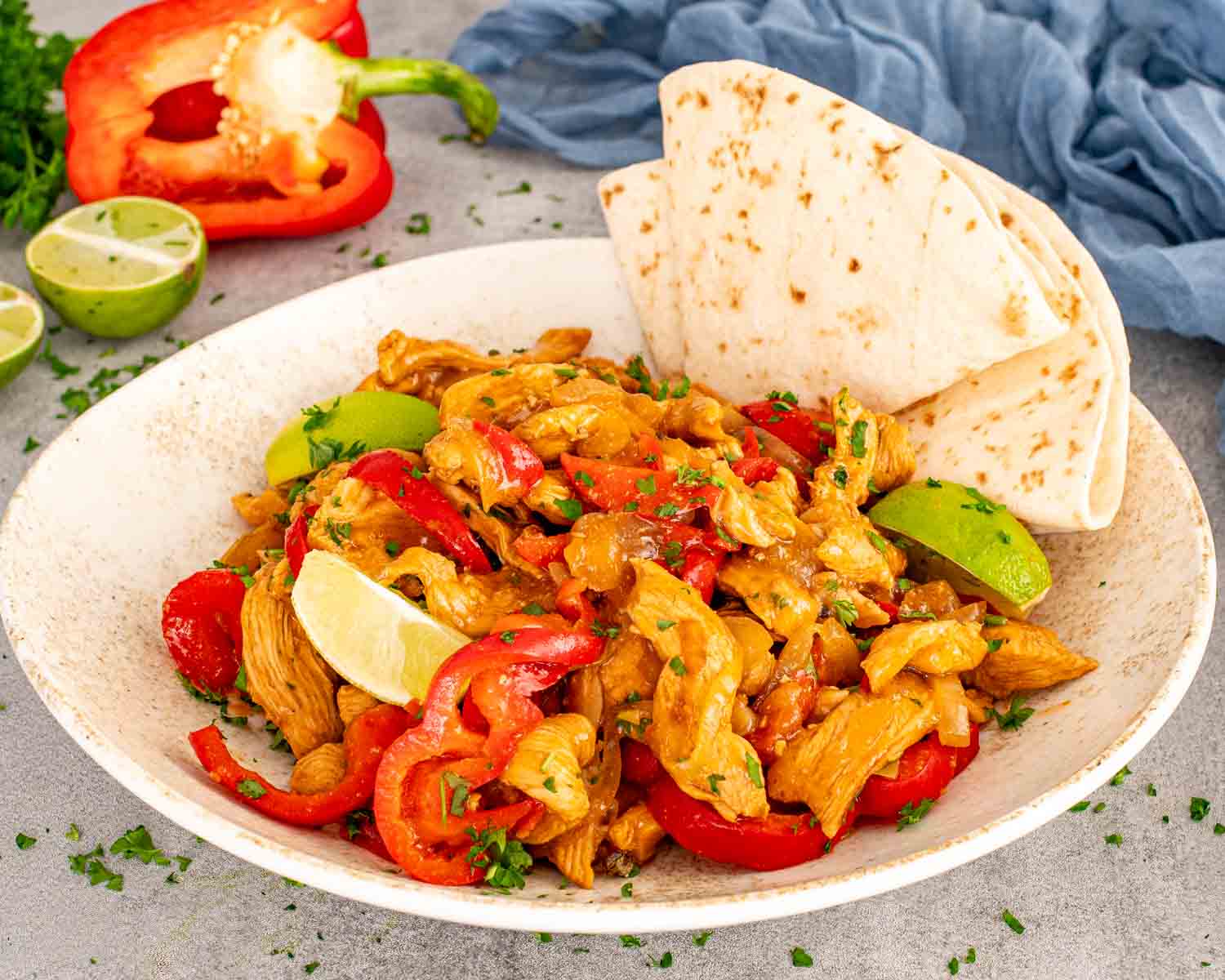 Platter of restaurant style chicken fajitas with red peppers, onions, lime wedges, fresh parsley, and warm folded tortillas.