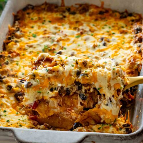 Close-up of a generous serving of cheesy Tex-Mex beef enchilada casserole lifted from a rustic metal baking dish.