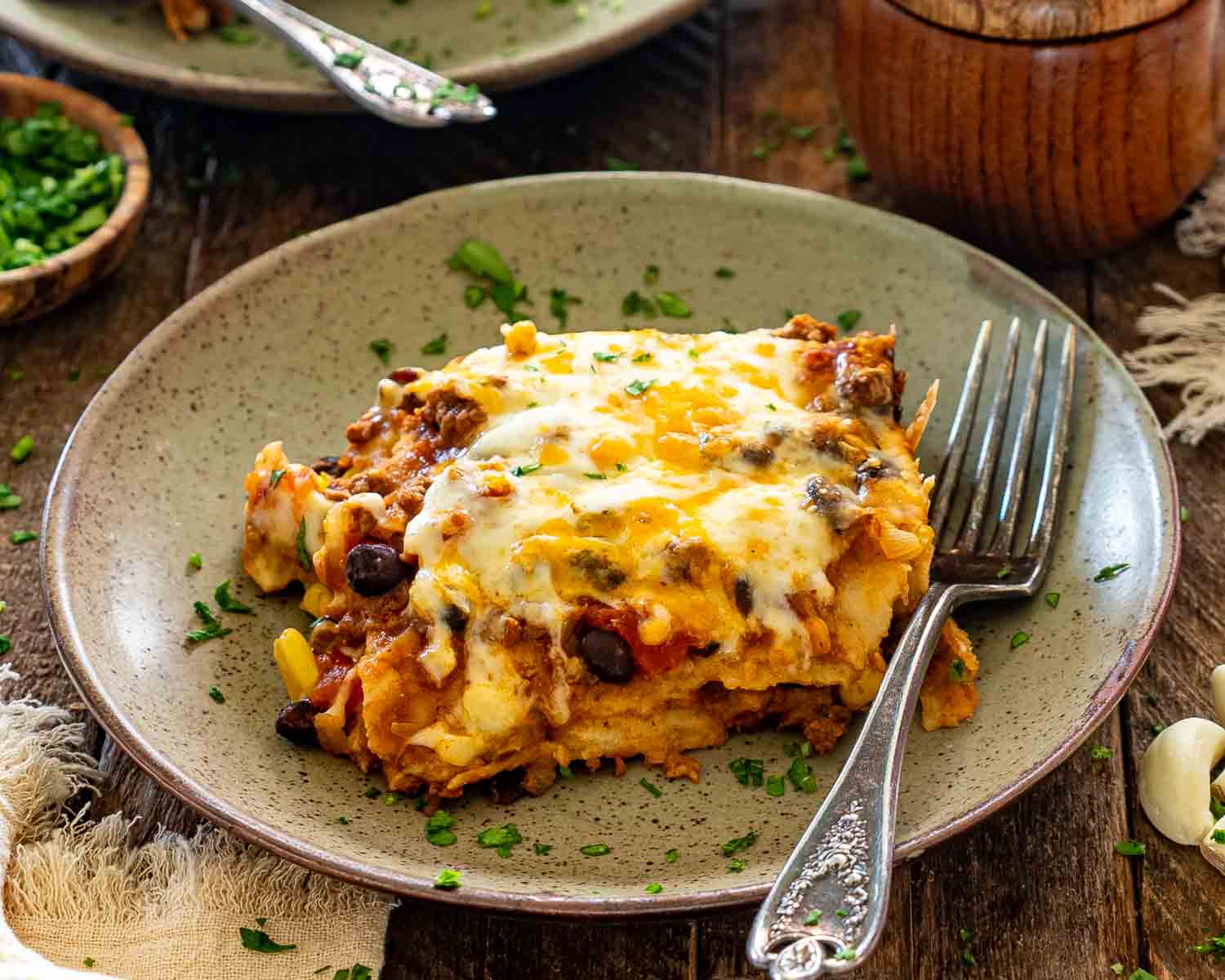 Cheesy slice of Tex-Mex beef enchilada casserole with black beans, corn, and tortillas served on a rustic plate.