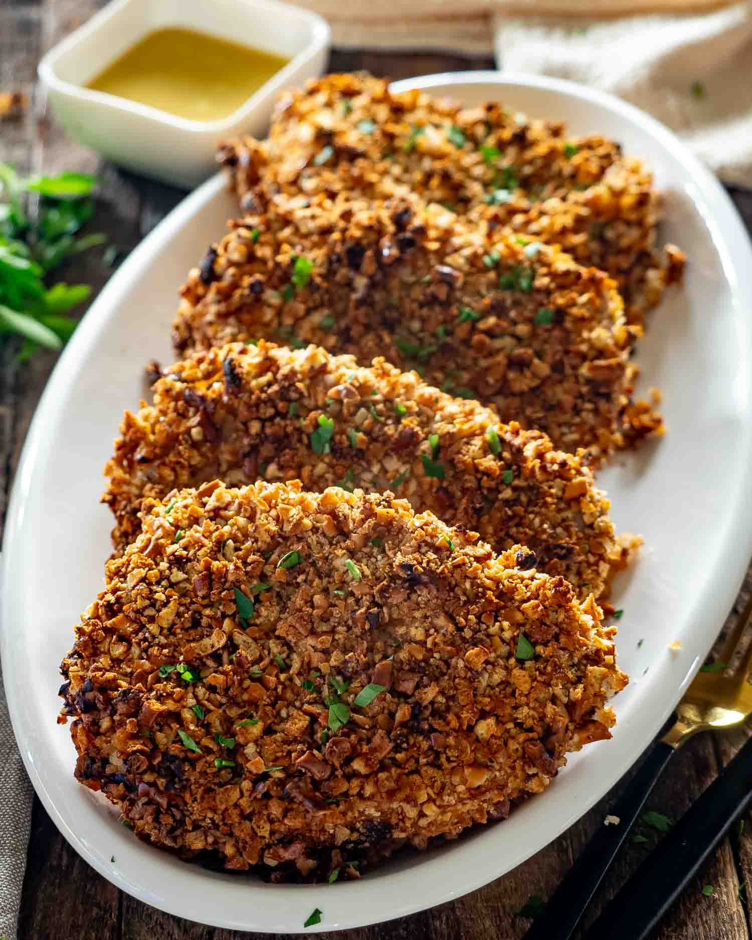 Four crispy pretzel-crusted pork chops garnished with parsley are arranged on a white platter with honey mustard sauce.
