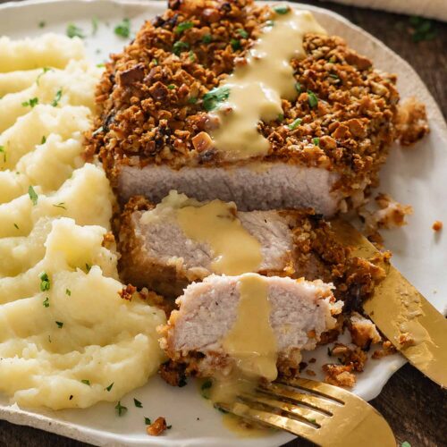 Sliced pretzel-crusted pork chops are served with mashed potatoes, honey mustard sauce, parsley, and gold cutlery on a plate.