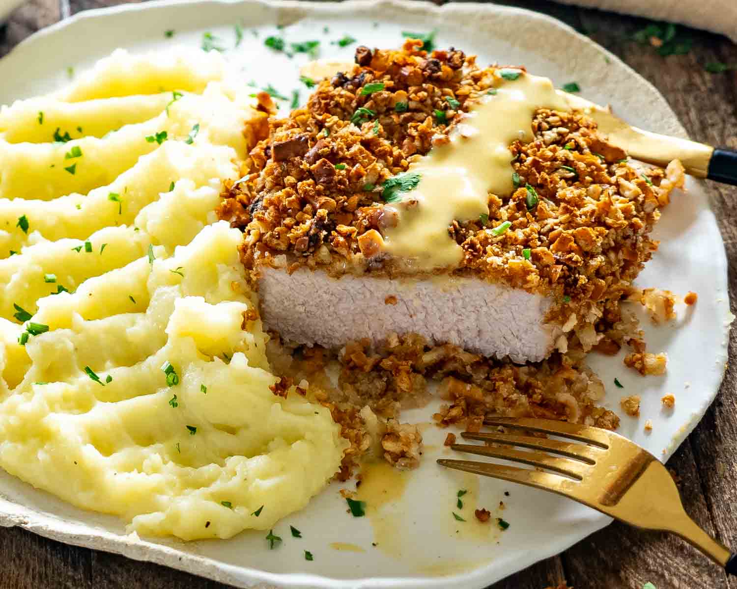 A sliced honey mustard pretzel-crusted pork chop rests beside parsley-flecked mashed potatoes with a gold fork in front.