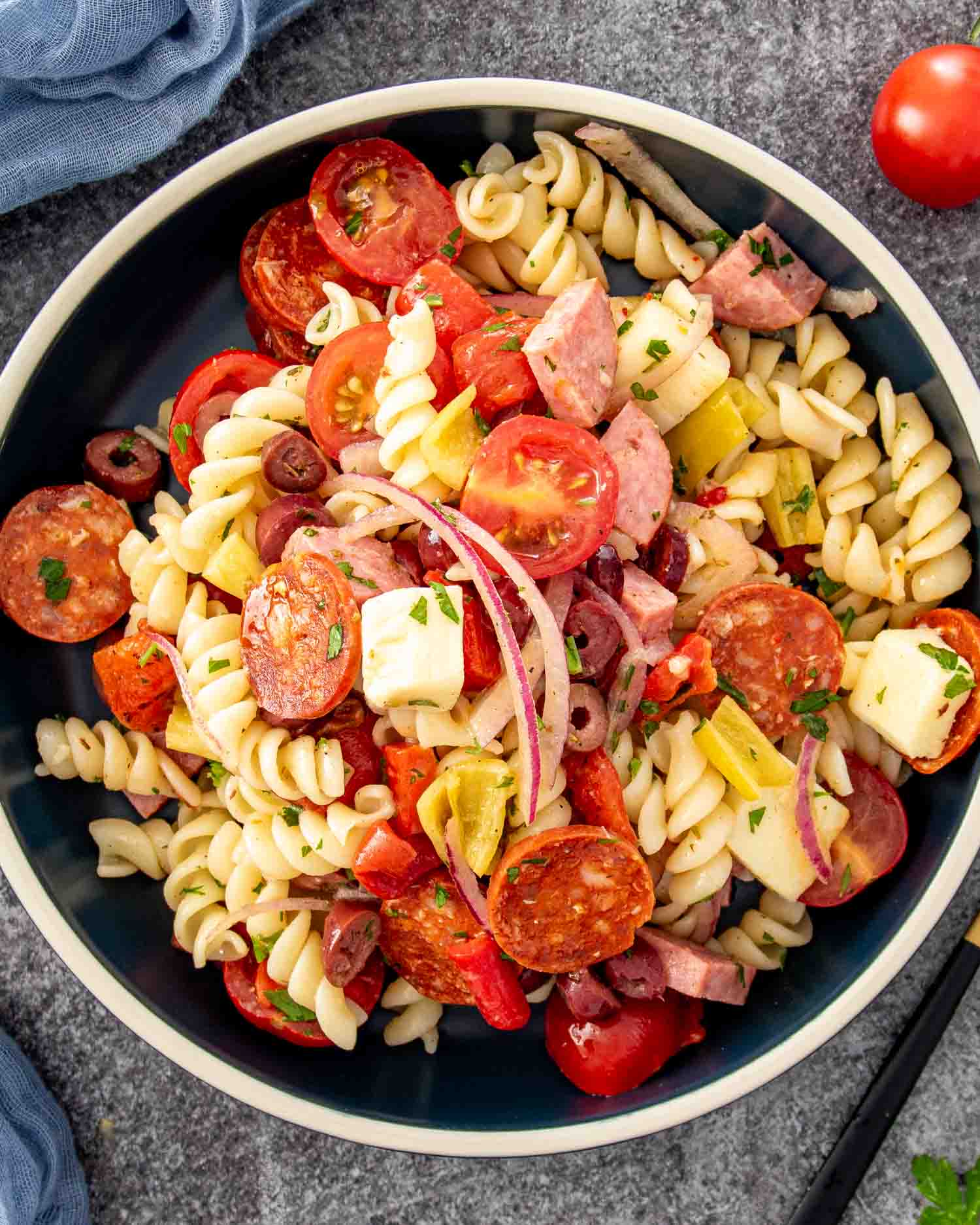 Overhead close-up of Italian pasta salad with rotini, pepperoni, salami, provolone, cherry tomatoes, olives, peperoncini, and red onion.