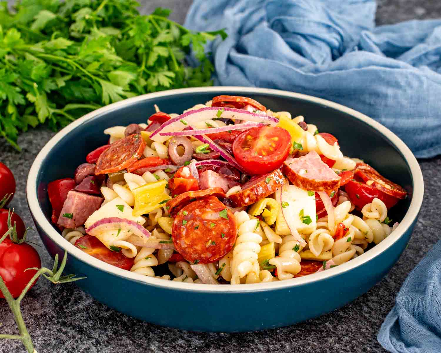 Italian pasta salad served in a dark bowl with pepperoni, salami, provolone, tomatoes, olives, red onion, and peperoncini.
