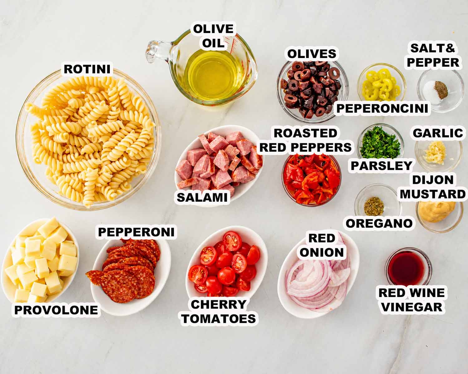 Overhead view of labeled Italian pasta salad ingredients, including rotini, meats, cheese, vegetables, olives, herbs, and homemade dressing ingredients.