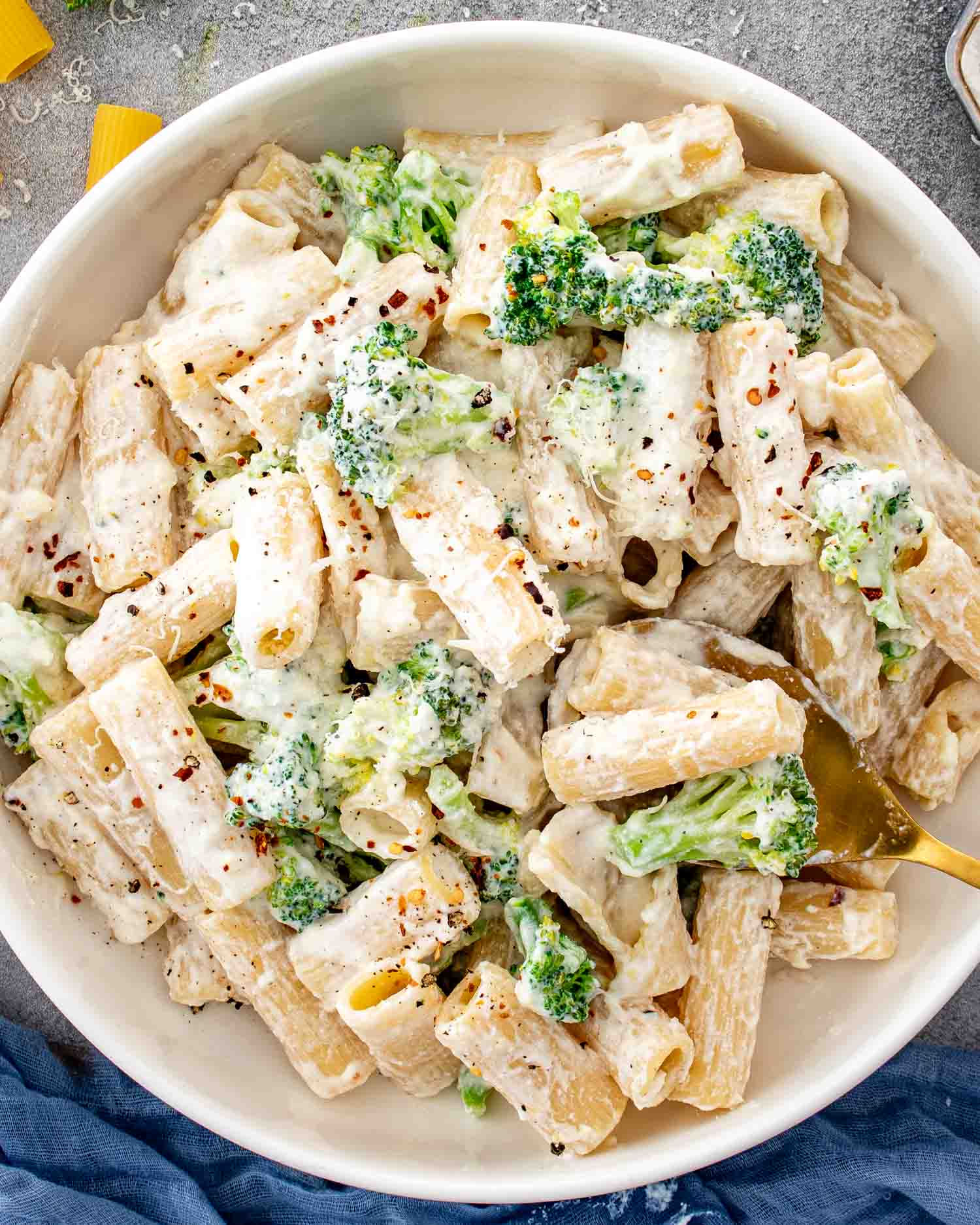 Overhead close-up of creamy lemon ricotta rigatoni with broccoli, Parmesan, red pepper flakes, and a gold serving spoon.