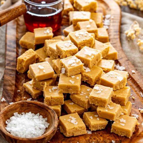 A generous pile of golden maple fudge squares sprinkled with flaky salt beside a glass of syrup.