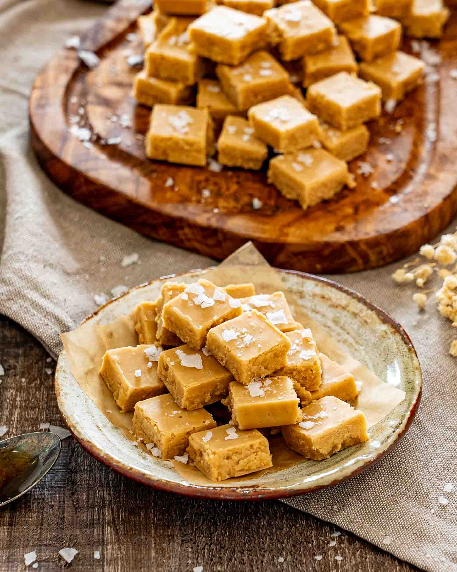 Salted maple fudge squares stacked on a rustic ceramic plate, with more pieces displayed behind on wood.