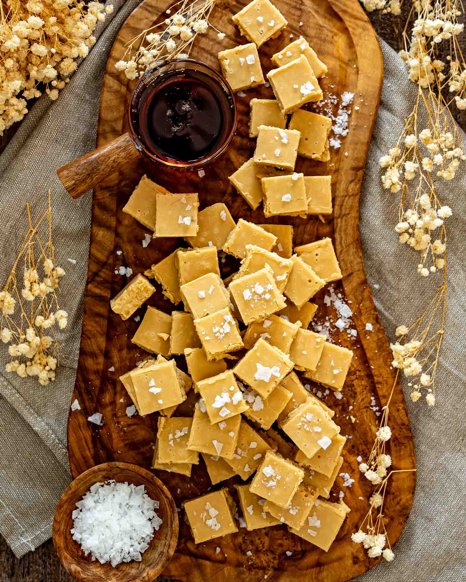 Overhead arrangement of salted maple fudge squares, maple syrup, and dried flowers styled across a long wooden board.