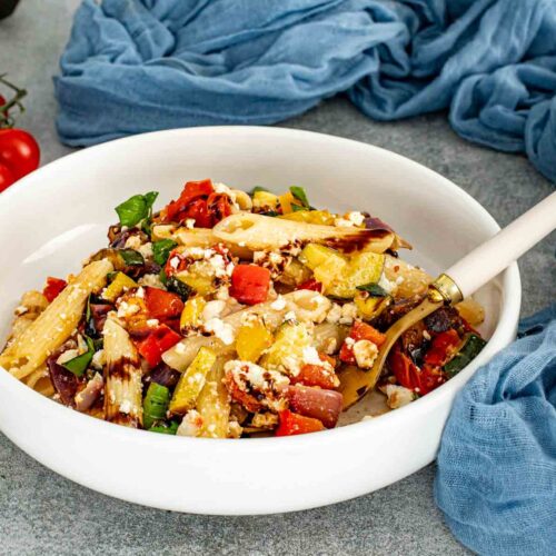 Roasted vegetable pasta salad with penne, feta, peppers, zucchini, tomatoes, and basil served in a shallow white bowl.