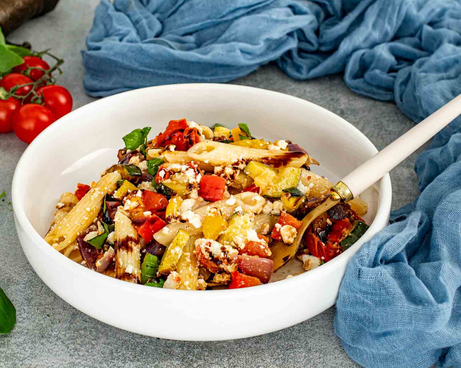 Roasted vegetable pasta salad with penne, feta, peppers, zucchini, tomatoes, and basil served in a shallow white bowl.