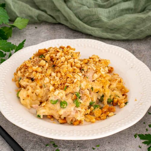 A generous serving of creamy tuna noodle casserole sits on a white plate with peas, parsley, and cracker crumbs.
