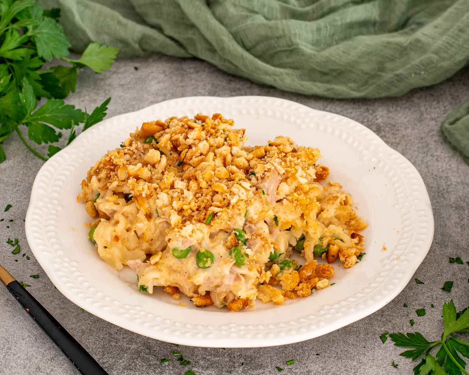 A generous serving of creamy tuna noodle casserole sits on a white plate with peas, parsley, and cracker crumbs.