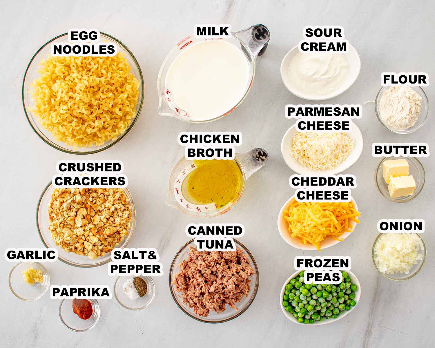 Overhead ingredient spread for tuna noodle casserole includes egg noodles, tuna, cheeses, peas, milk, seasonings, butter, and crackers.