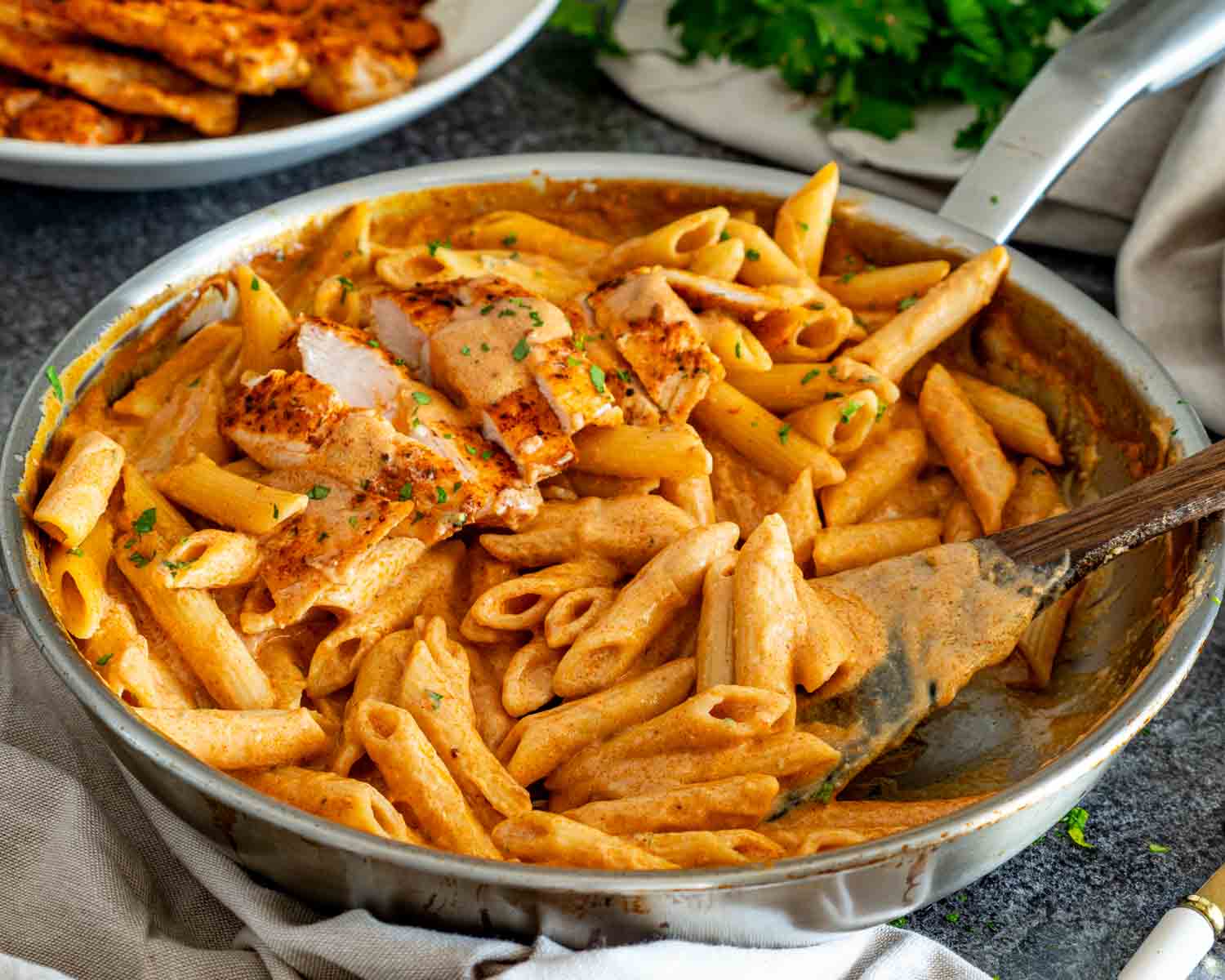 Serving spoon scoops creamy garlic Parmesan penne from a skillet topped with sliced chicken and fresh chopped parsley.
