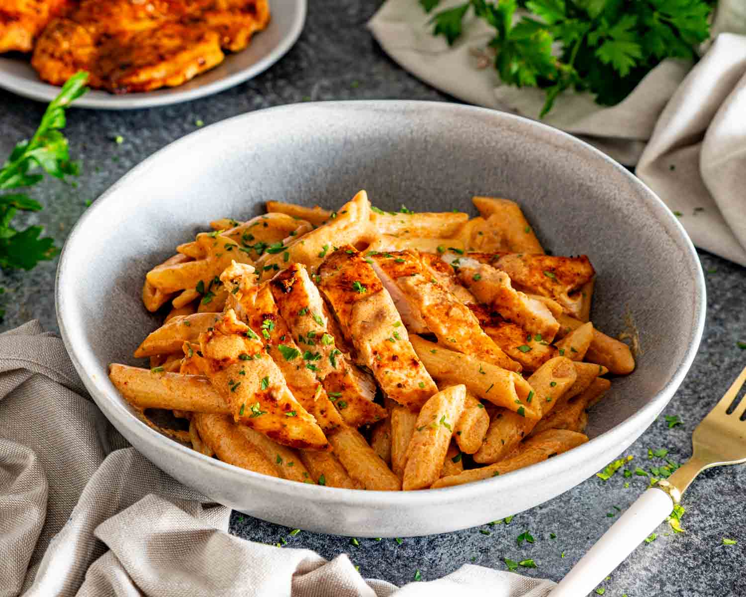 Bowl of creamy garlic Parmesan chicken pasta topped with sliced golden chicken and parsley beside the serving skillet.