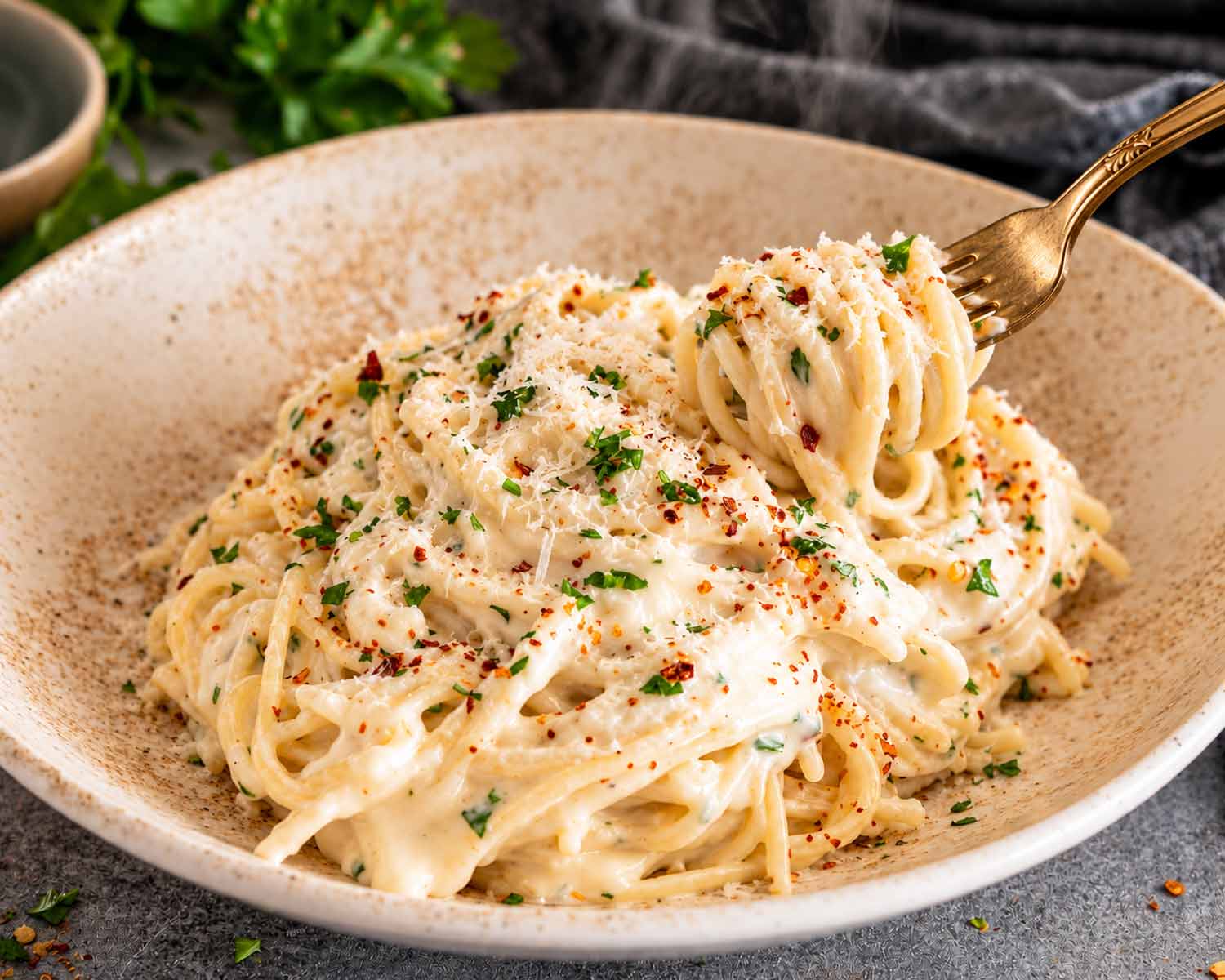 Creamy Parmesan spaghetti is twirled around a gold fork above a rustic bowl garnished with parsley and red pepper flakes.