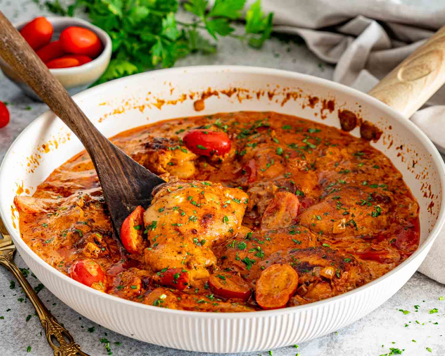 Creamy sun-dried tomato chicken thighs simmering in a white skillet with tomatoes, herbs, and a wooden spoon.