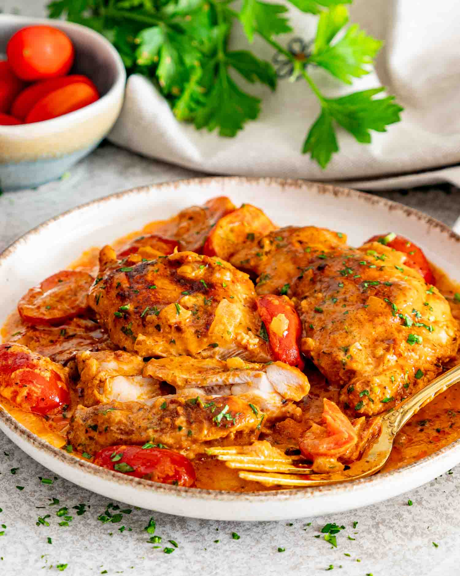 Close-up of sliced creamy sun-dried tomato chicken thighs with cherry tomatoes, herbs, and a golden fork.