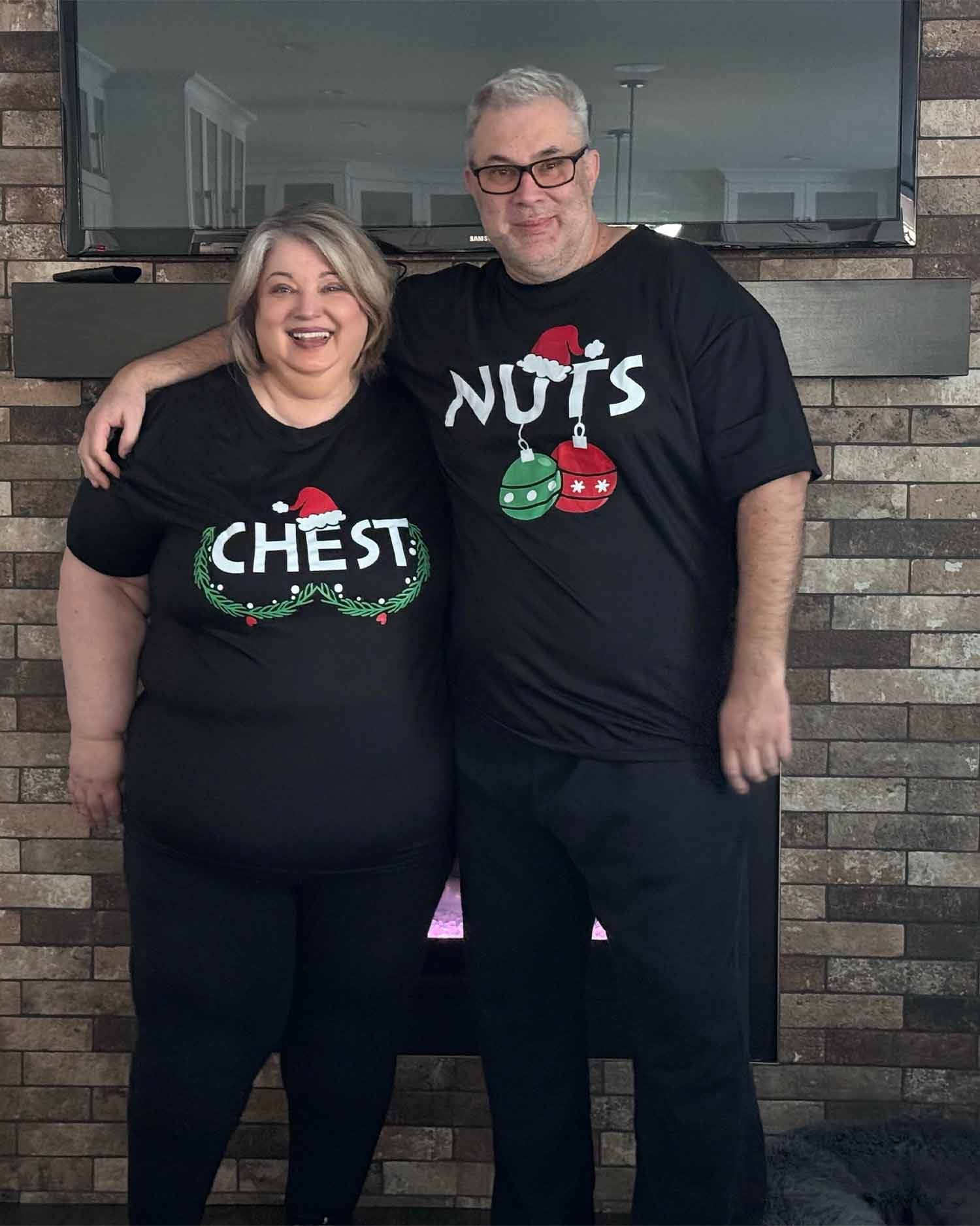 Joanna Cismaru and her husband Remo smiling together in matching funny Christmas shirts while standing in front of their fireplace.