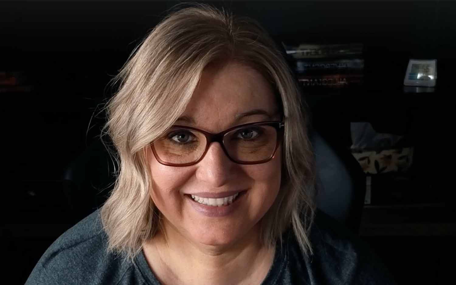 Joanna Cismaru smiling at the camera wearing glasses, with shoulder-length blonde hair and a dark gray top.