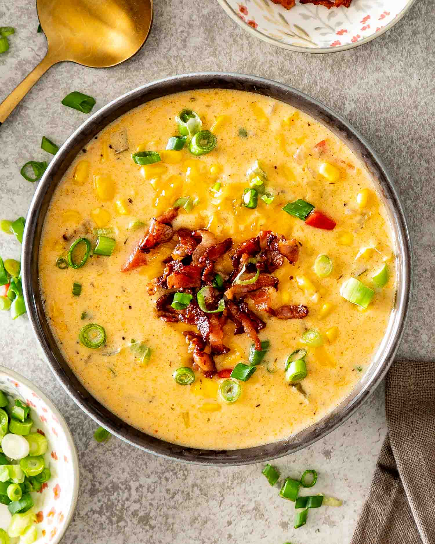 Overhead bowl of creamy loaded corn chowder topped with bacon, cheddar cheese, and green onions beside a gold spoon.