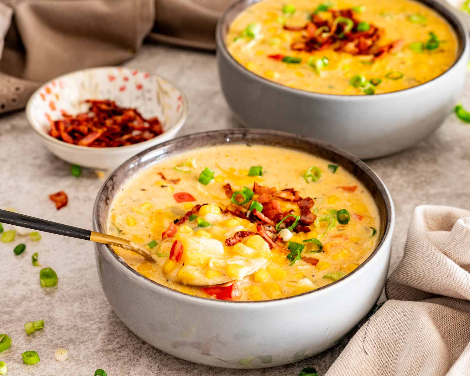 Spoon lifting a creamy bite of loaded corn chowder with potatoes, corn, bacon, cheddar, peppers, and green onions.
