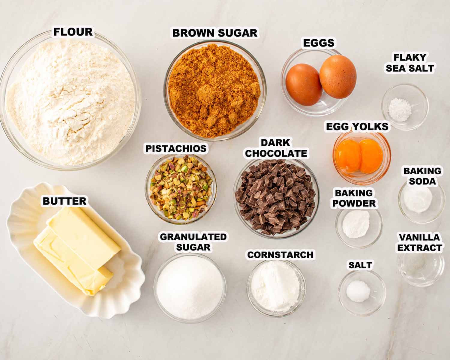 Labeled bowls of flour, sugars, eggs, butter, pistachios, dark chocolate and baking ingredients arranged on a light surface.