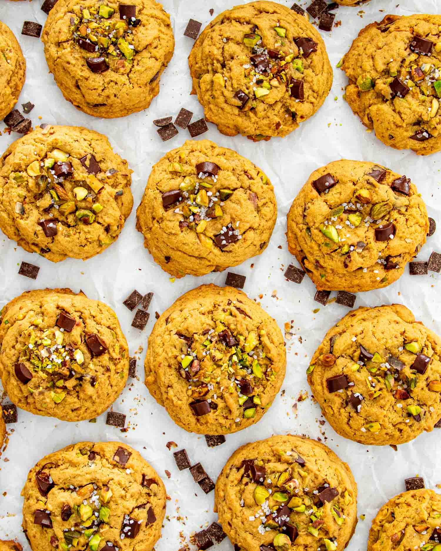 Overhead view of thick pistachio chocolate chunk cookies sprinkled with flaky sea salt and surrounded by scattered chocolate pieces.