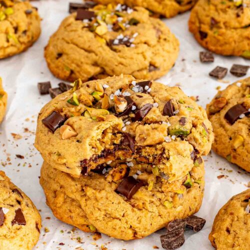 Broken pistachio chocolate chunk cookies reveal gooey melted chocolate among whole cookies scattered with pistachios and chocolate pieces.