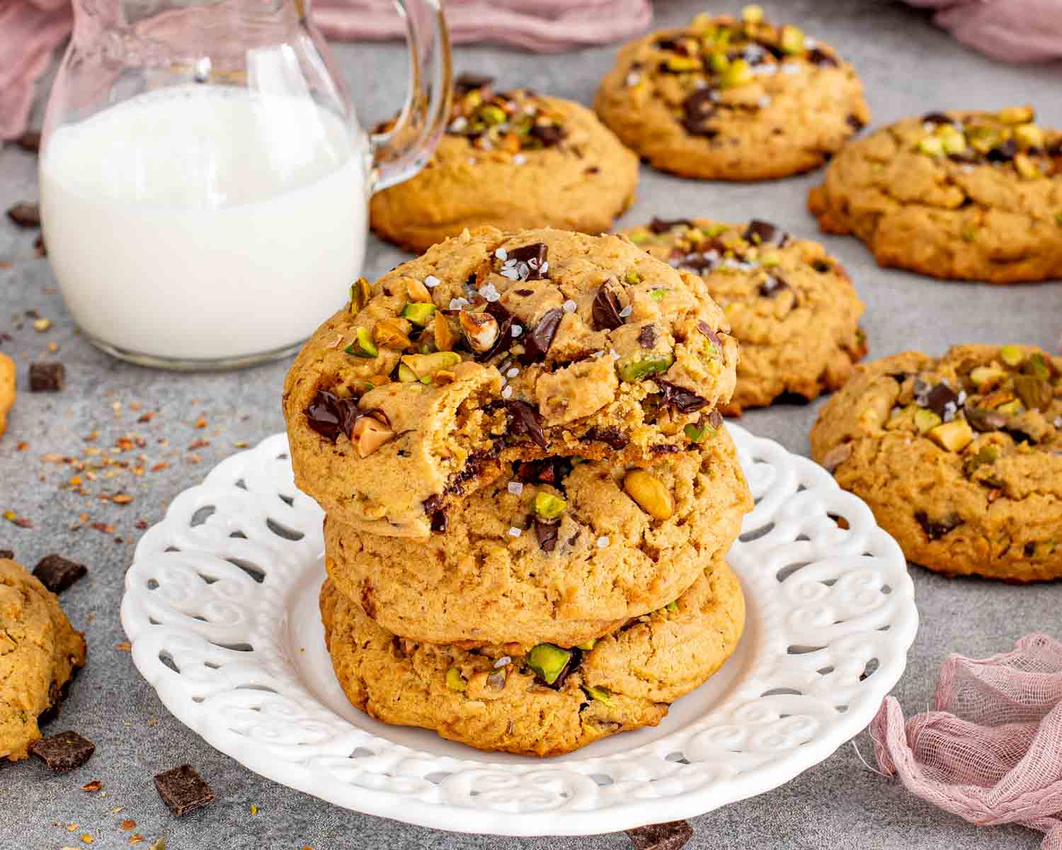 Three thick pistachio chocolate chunk cookies are stacked on a white plate, topped with a broken chocolate-filled cookie.