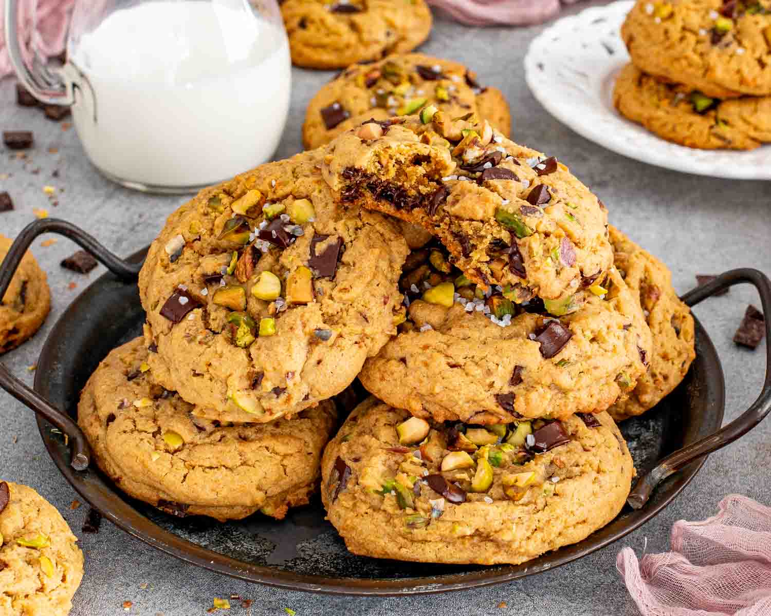 A broken pistachio chocolate chunk cookie rests atop a generous pile of thick cookies beside a glass of milk.