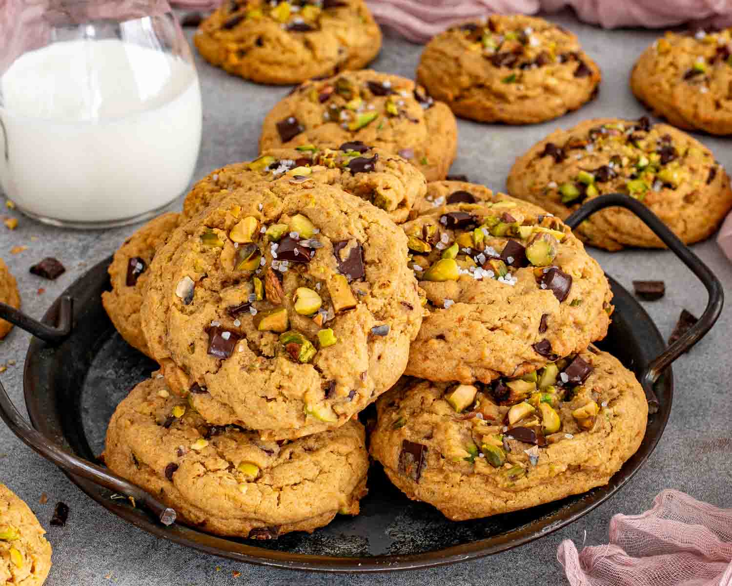 Thick pistachio chocolate chunk cookies piled on a dark serving tray with a glass of milk in the background.